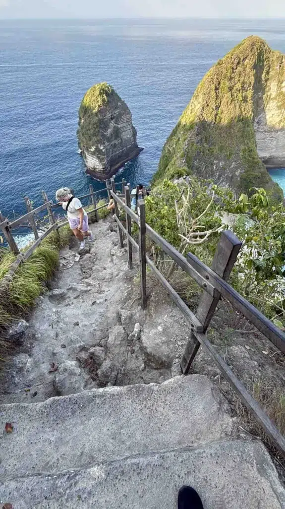 Vue plongeante sur les marches usées de la descente de Kelingking Beach, avec un garde-fou en bois, une randonneuse qui remonte et les rochers en mer en contrebas